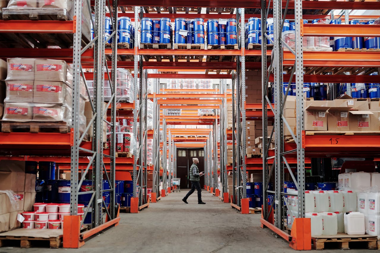services-04 A man walking through a large industrial warehouse with stacked shelves filled with goods and products.